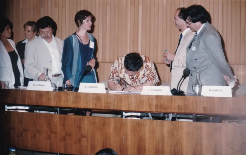 A signatory signs the Charter at the official table, surrounded by standing delegates.