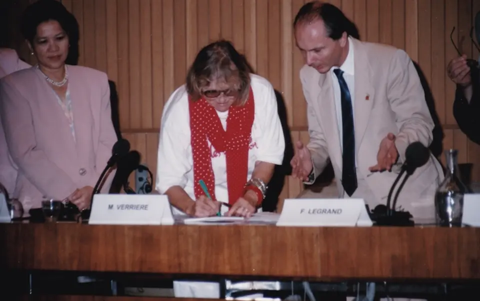 A signatory wearing a red scarf signs the Charter; a standing man points to the page.