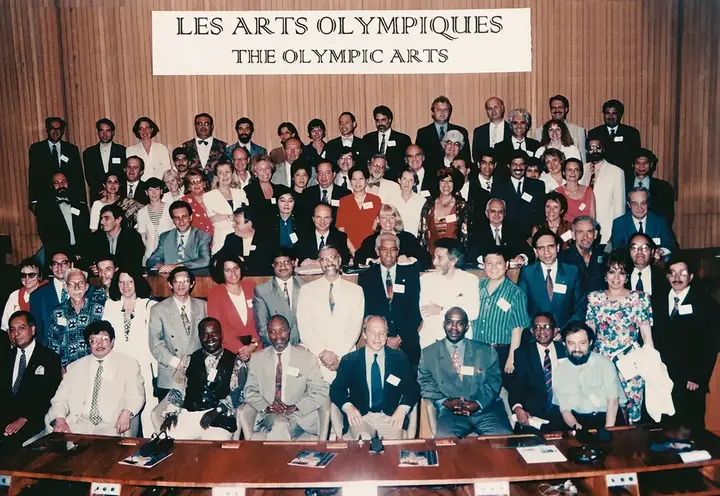 About a hundred people pose in rows in a UNESCO hall, beneath a banner reading “Les Arts Olympiques – The Olympic Arts”.